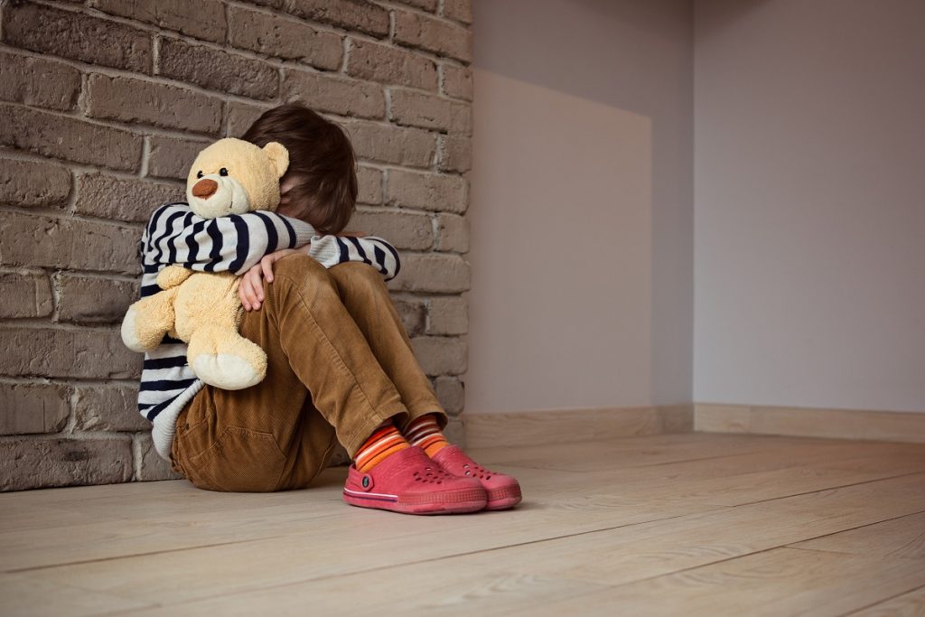 Sad little boy sitting against the wall in despair. In his hands he holds an old friend teddy bear