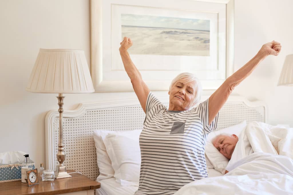 Senior woman sitting on her bed in the morning yawning with arms raised in a stretch