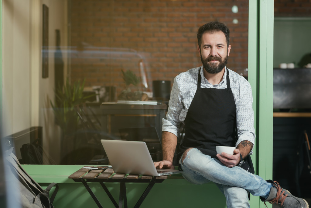 Entrepreneur sitting on a ledge while checking his laptop and holding a small white cup.