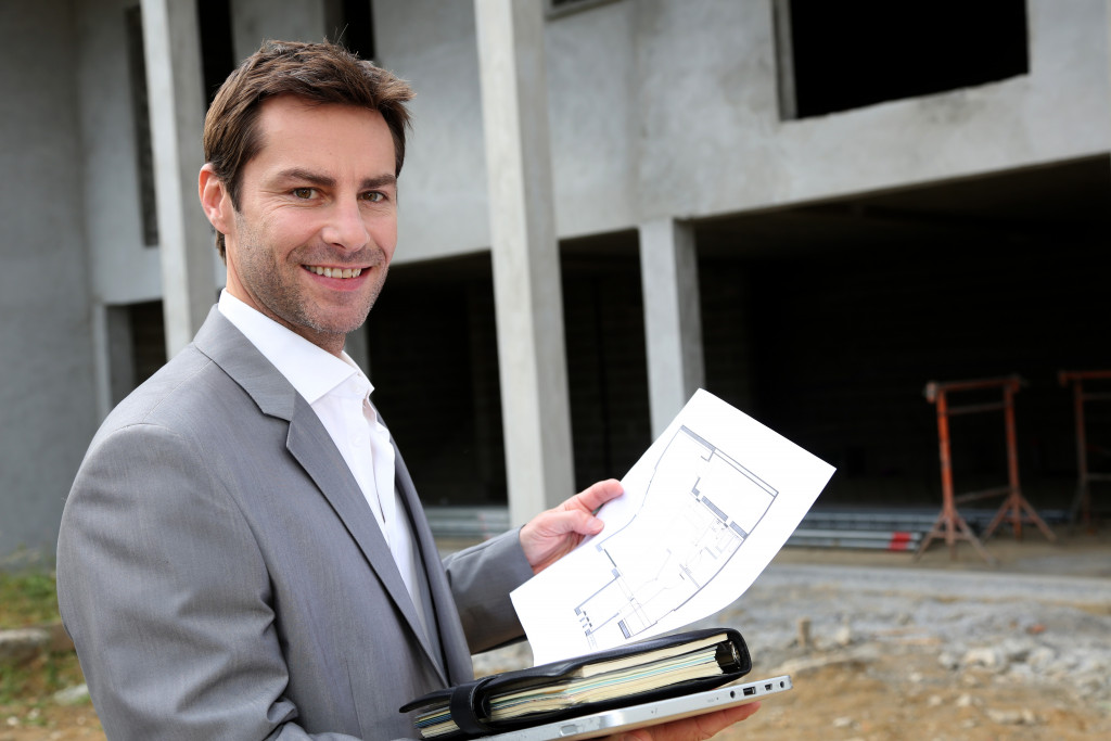 A man  in a suit holding a floor plan of the under-construction building behind him